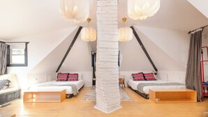 A modern loft bedroom with a sloped ceiling, two beds with patterned pillows, a central white column, and pendant lighting.