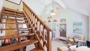 A spacious living area with a wooden staircase, a ceiling fan, and a view of the outdoors through large windows.