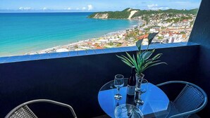 A balcony with a glass table and two chairs, overlooking a beach and a town.