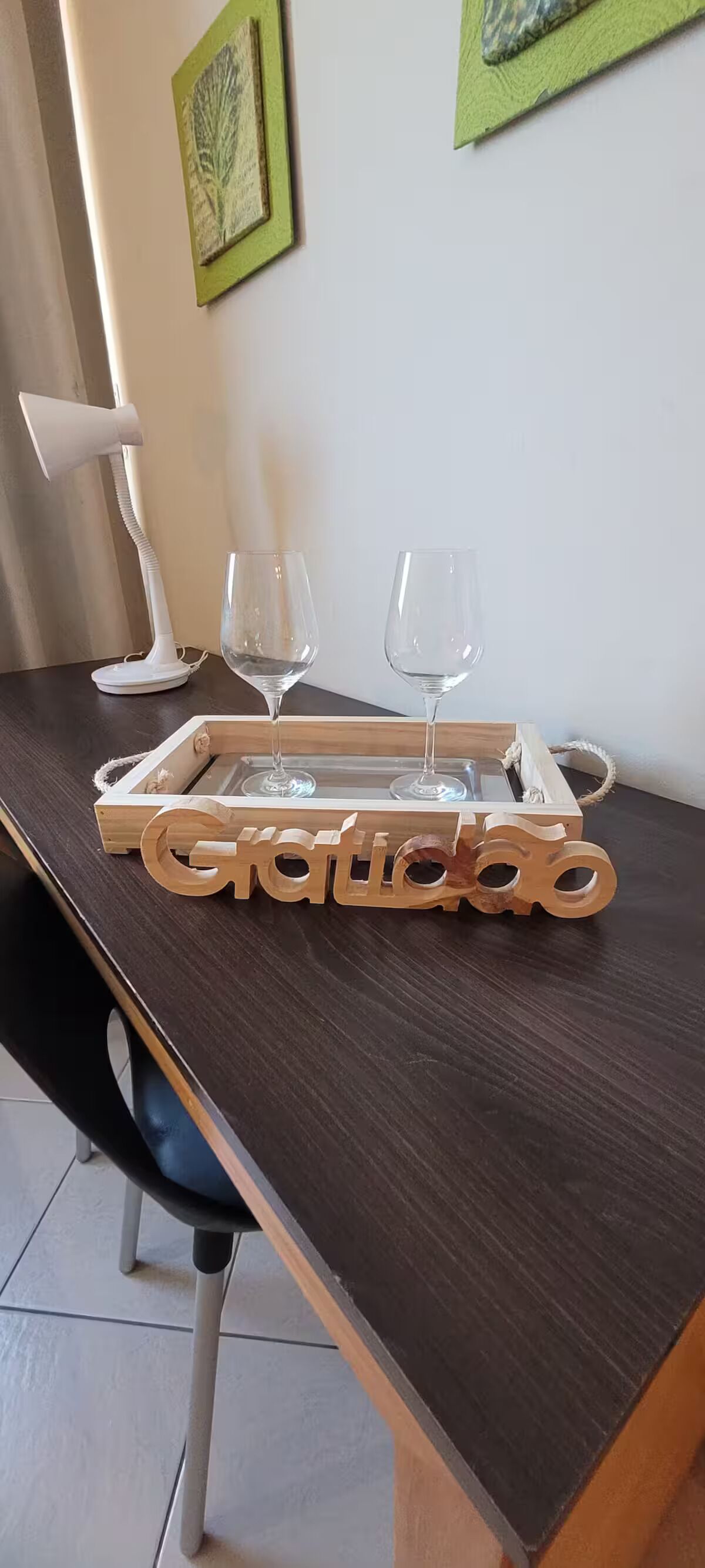 Un plateau en bois sur lequel est inscrit le mot « Gratitude », posé sur une table en bois foncé, accompagné de deux verres à vin et d’une lampe blanche.