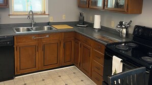 Une cuisine avec des meubles de rangement en bois, un poêle noir, un évier et une fenêtre avec des stores.
