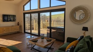 Uma sala de estar ampla com uma grande porta de vidro deslizante para um deck ao ar livre, teto de madeira e um sofá verde com almofadas estampadas.