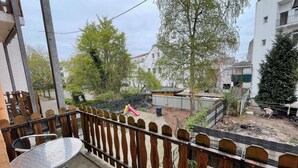 Een balkon met een metalen tafel en stoelen, een houten hek en uitzicht op een woongebied met bomen en gebouwen.