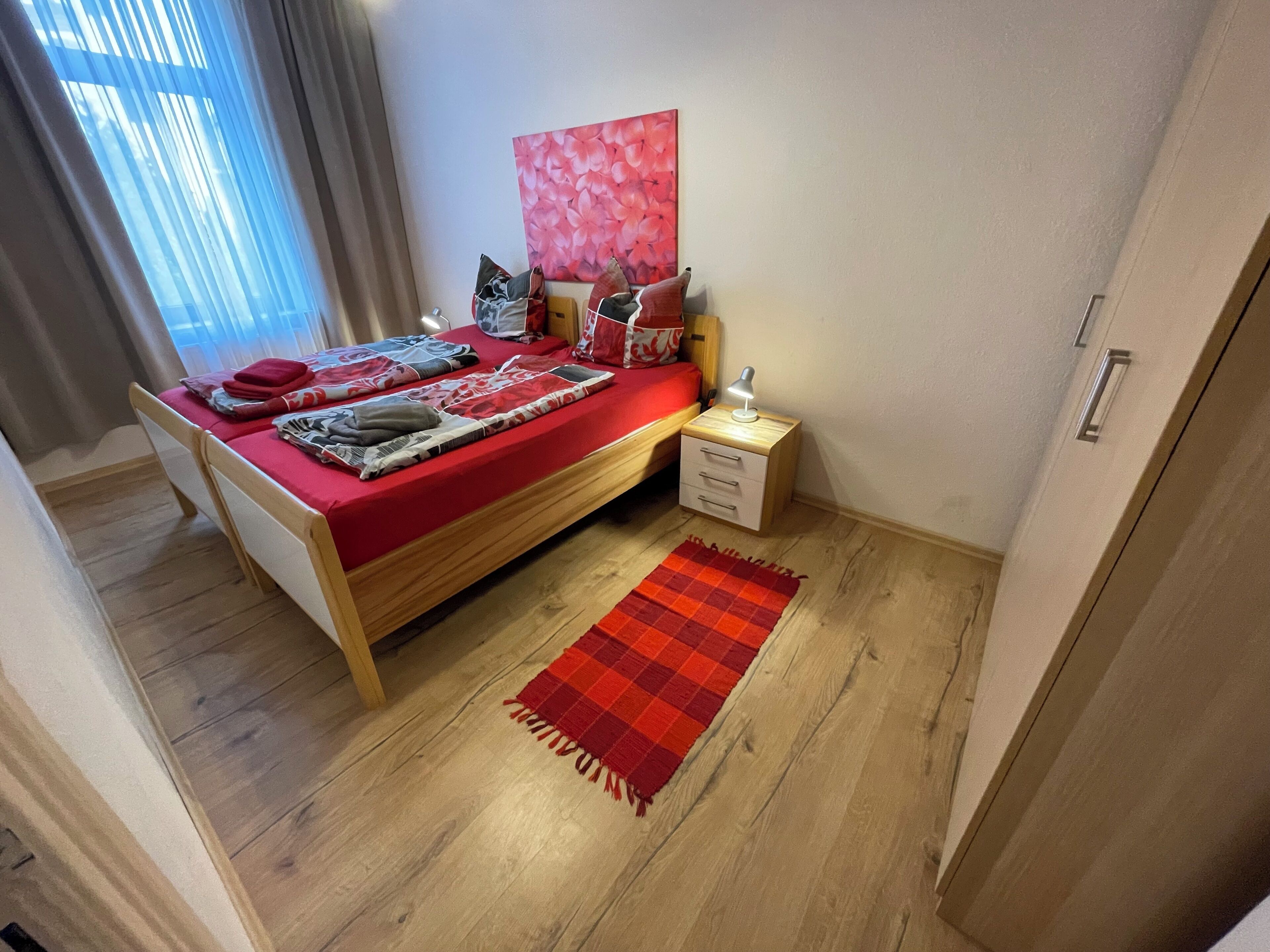 A neatly arranged bedroom with a wooden bed, a red blanket, and a small bedside table with a lamp.