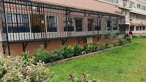 A building with a long balcony with black metal railings, a grassy area with plants, and a person in the background. - SS LUMINA GUEST HOUSE (Bengaluru)