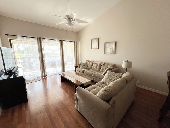 Ein Wohnzimmer mit einem beigen Sofa, einem Couchtisch, einem Fernseher und einem Deckenventilator.
