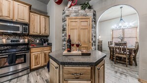 A kitchen with wooden cabinets, a granite countertop, a stone accent wall, and a dining area with a chandelier. - Toasted Marshmallows | Luxury Cabin in Ruidoso w/ Hot Tub & Game Room (Ruidoso)