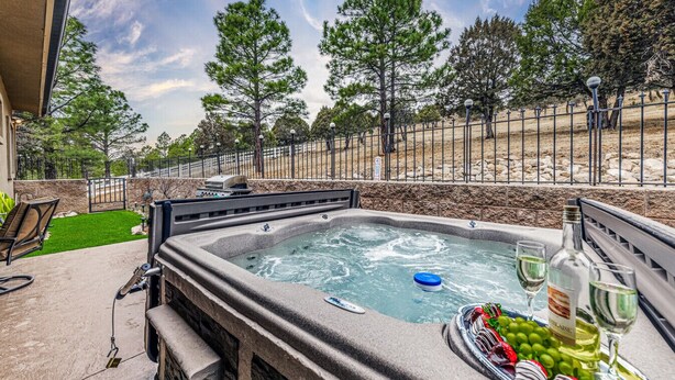 A hot tub with a stone surround, a patio chair, and a table with a bottle of wine and grapes. - Toasted Marshmallows | Luxury Cabin in Ruidoso w/ Hot Tub & Game Room (Ruidoso)