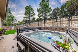 A hot tub with a stone surround, a patio chair, and a table with a bottle of wine and grapes. - Toasted Marshmallows | Luxury Cabin in Ruidoso w/ Hot Tub & Game Room (Ruidoso)