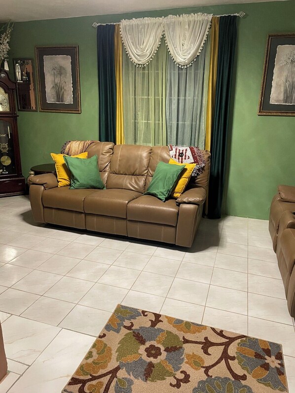 A living room with a brown leather sofa, green and yellow cushions, a patterned rug, and a green wall with framed artwork.