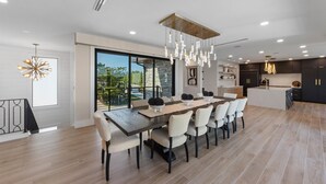 A modern dining area with a long table, multiple chairs, and a chandelier. - Lido Luxe Estate: Large Rental Steps to the Beach with Pool, Spa, Outdoor Games (Lido Key)