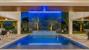 A swimming pool with a waterfall feature, surrounded by lounge chairs and palm trees. - Lido Luxe Estate: Large Rental Steps to the Beach with Pool, Spa, Outdoor Games (Lido Key)