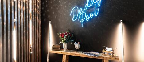 A room with a neon sign reading "Buddy Pool," a wooden table with books and a vase of flowers, and a shovelful of snow on the floor.