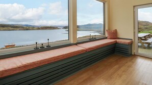 Habitación con un ventanal grande que da a un lago y colinas, un banco con cojines y suelo de madera.