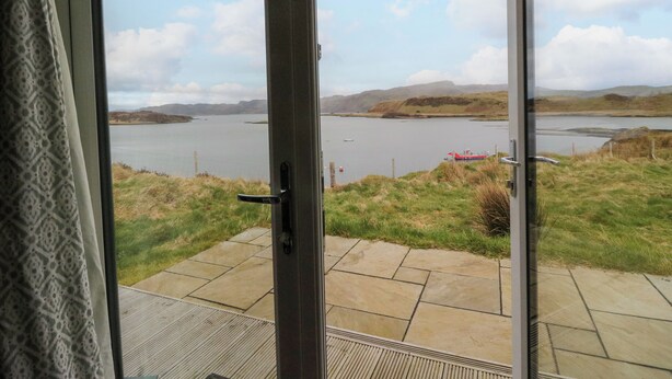 A view from a room with a glass door leading to a patio overlooking a lake with hills in the distance. - Creags (Oban)