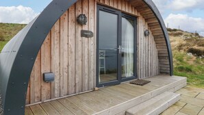 A wooden cabin with a curved metal roof, a sliding glass door, and a small outdoor seating area with steps leading up to the entrance. - Creags (Oban)