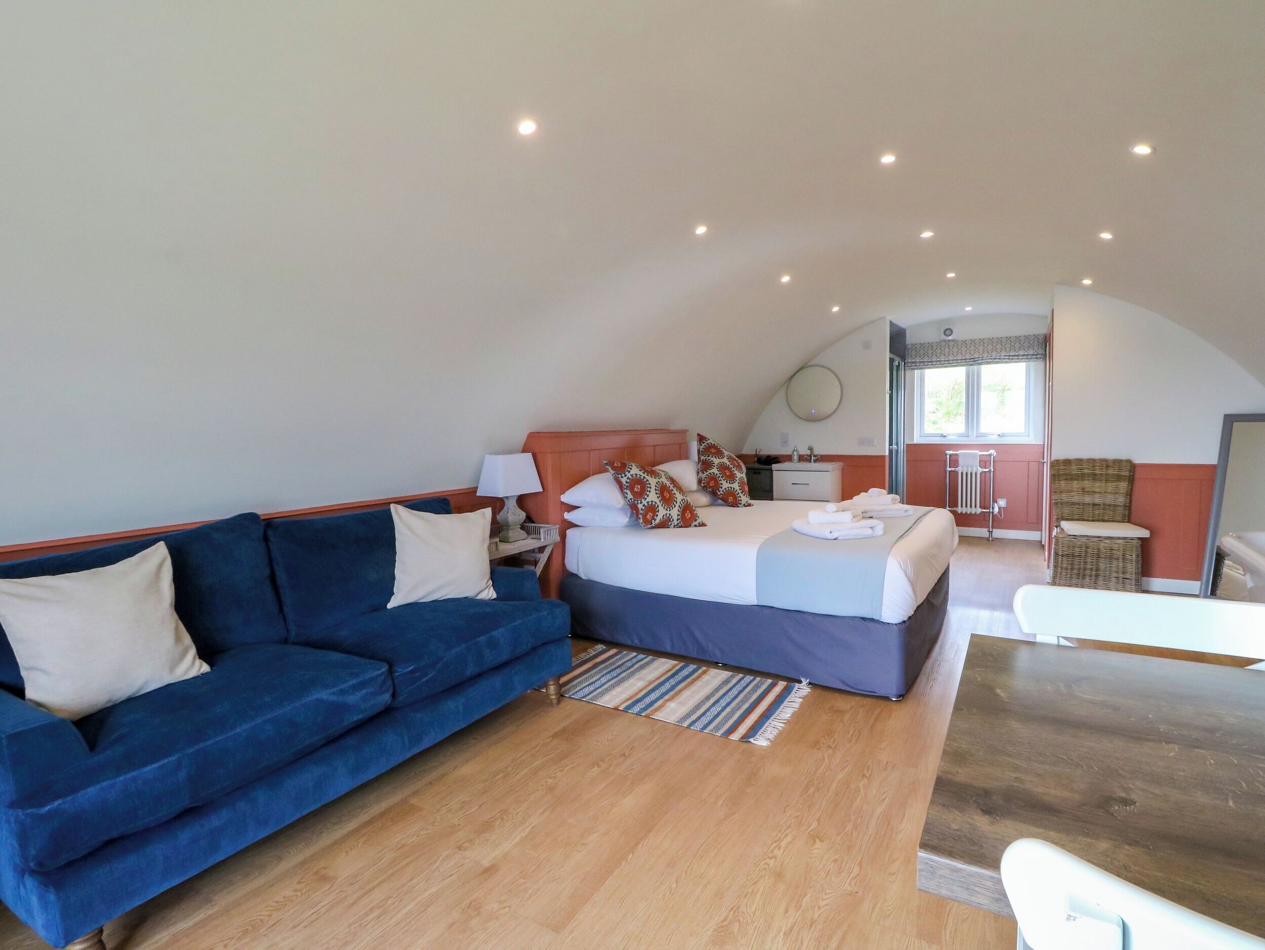 Une chambre à coucher avec un lit, un canapé bleu, un plancher en bois, une petite table et une salle de bain visible à travers une porte ouverte.