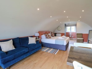 A bedroom with a bed, a blue sofa, a wooden floor, a small table, and a bathroom visible through an open door. - Fladda (Oban)