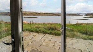 Uitzicht vanuit een kamer met een glazen deur naar een terras, met uitzicht op een meer met grasoevers en heuvels in de verte.