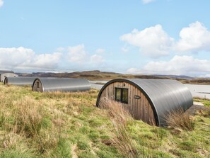 A series of dome-shaped cabins with a wooden exterior and metal roof, situated in a grassy field with a lake and hills in the background. - Insh (Kingussie)