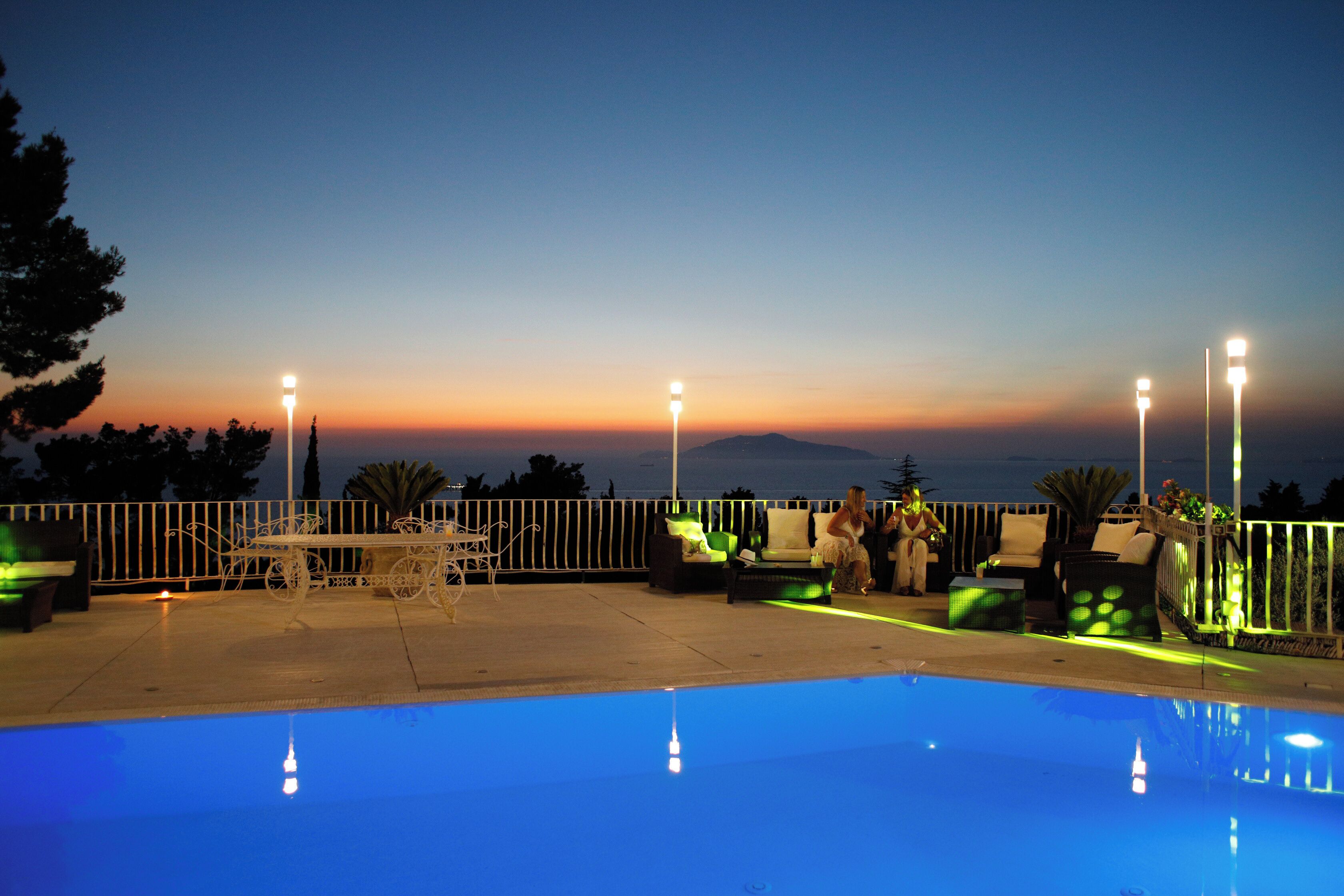 An outdoor pool area with seating and lighting, overlooking a sunset.