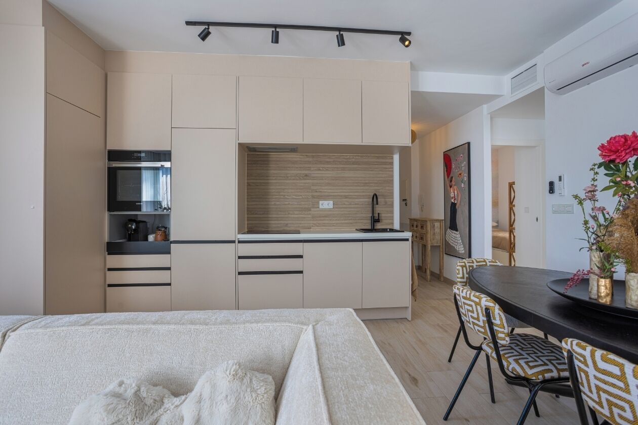 A modern kitchen with built-in appliances, a dining area with patterned chairs, and a bed with a white cover.