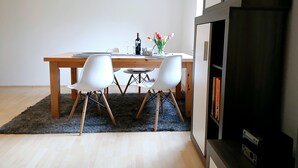 Un comedor con una mesa de madera, dos sillas blancas, un jarrón pequeño con flores y una botella de vino.
