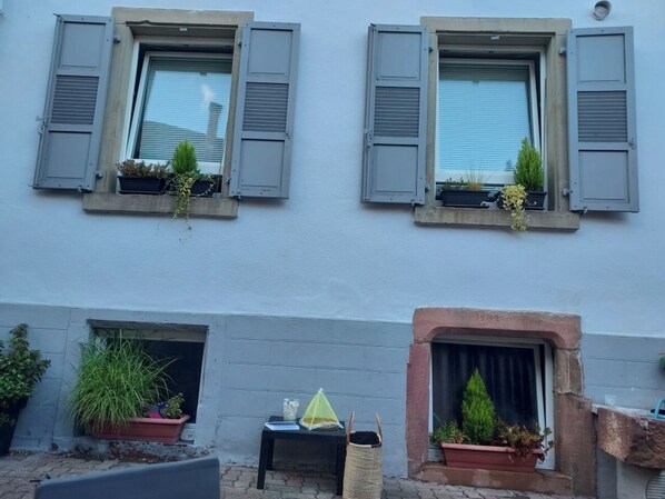 A building with two windows, each with shutters, and a small brick fireplace below.