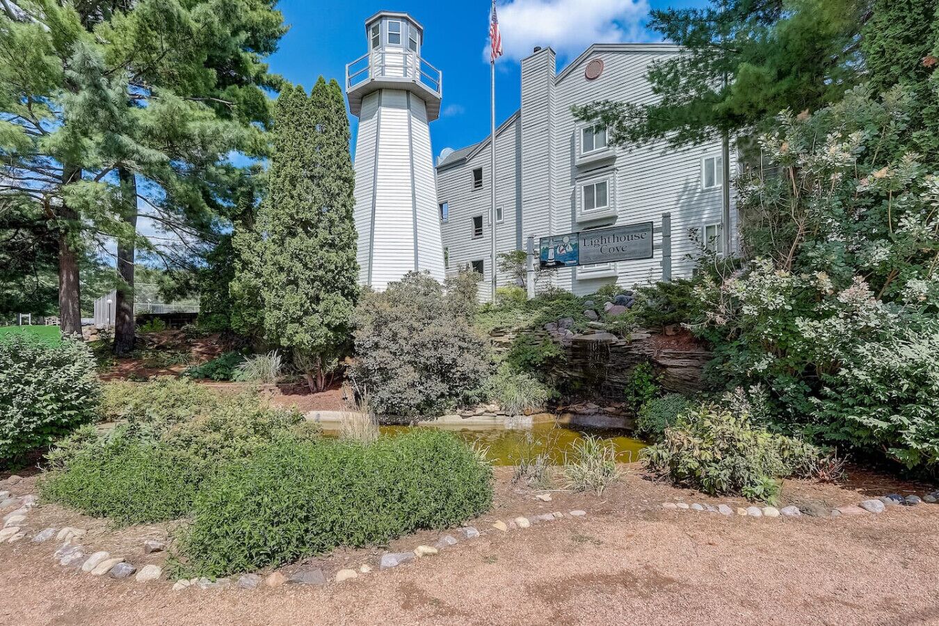 A historic building with a lighthouse tower, surrounded by a well-maintained garden with a pond and various shrubs.