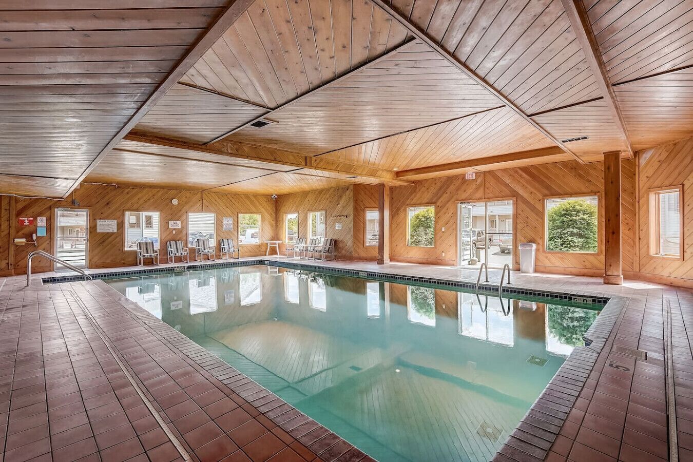 A large indoor swimming pool with wooden walls and ceiling, surrounded by lounge chairs and a tiled floor.