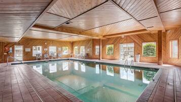 A large indoor swimming pool with wooden walls and ceiling, surrounded by lounge chairs and a tiled floor.