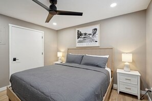 A modern bedroom with a bed, a ceiling fan, a framed picture of a river, and bedside lamps.