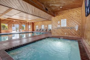 A large indoor swimming pool with wooden walls and ceiling, clear glass doors leading outside, and a tiled floor.