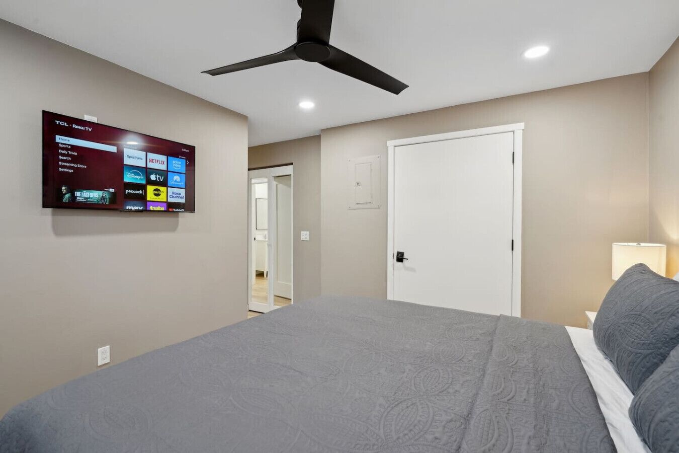 A bedroom with a bed, a ceiling fan, a TV mounted on the wall displaying streaming services, and a door leading to another room.