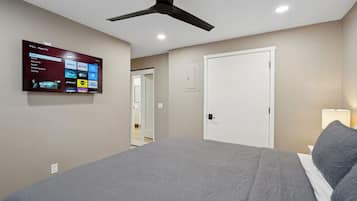 A bedroom with a bed, a ceiling fan, a TV mounted on the wall displaying streaming services, and a door leading to another room.