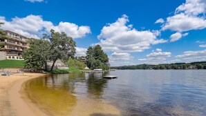 A lakeside view with a sandy beach, a dock, and a building with multiple balconies. - Pine Needle 506 at Lighthouse Cove (Wisconsin Dells)
