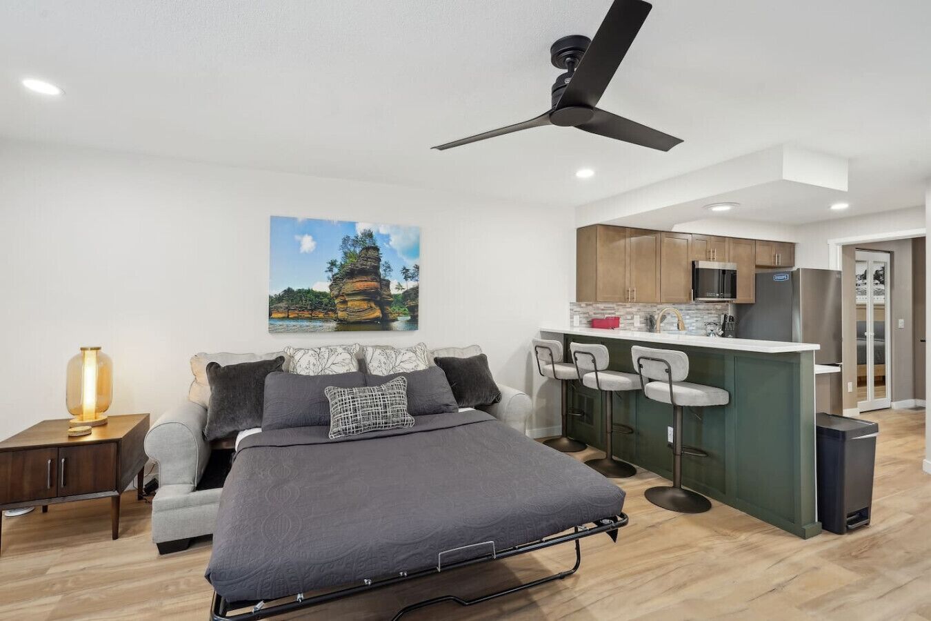 A modern bedroom with a bed, a desk, a chair, a nightstand, a ceiling fan, a kitchen area with a bar, and a dining area.