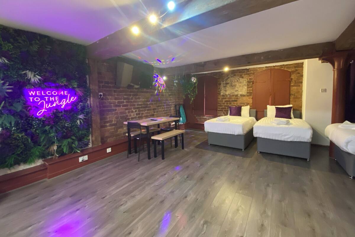 Una habitación con pared de ladrillo, un letrero de neón que dice "Welcome to the Jungle", una mesa de madera con sillas y dos camas.