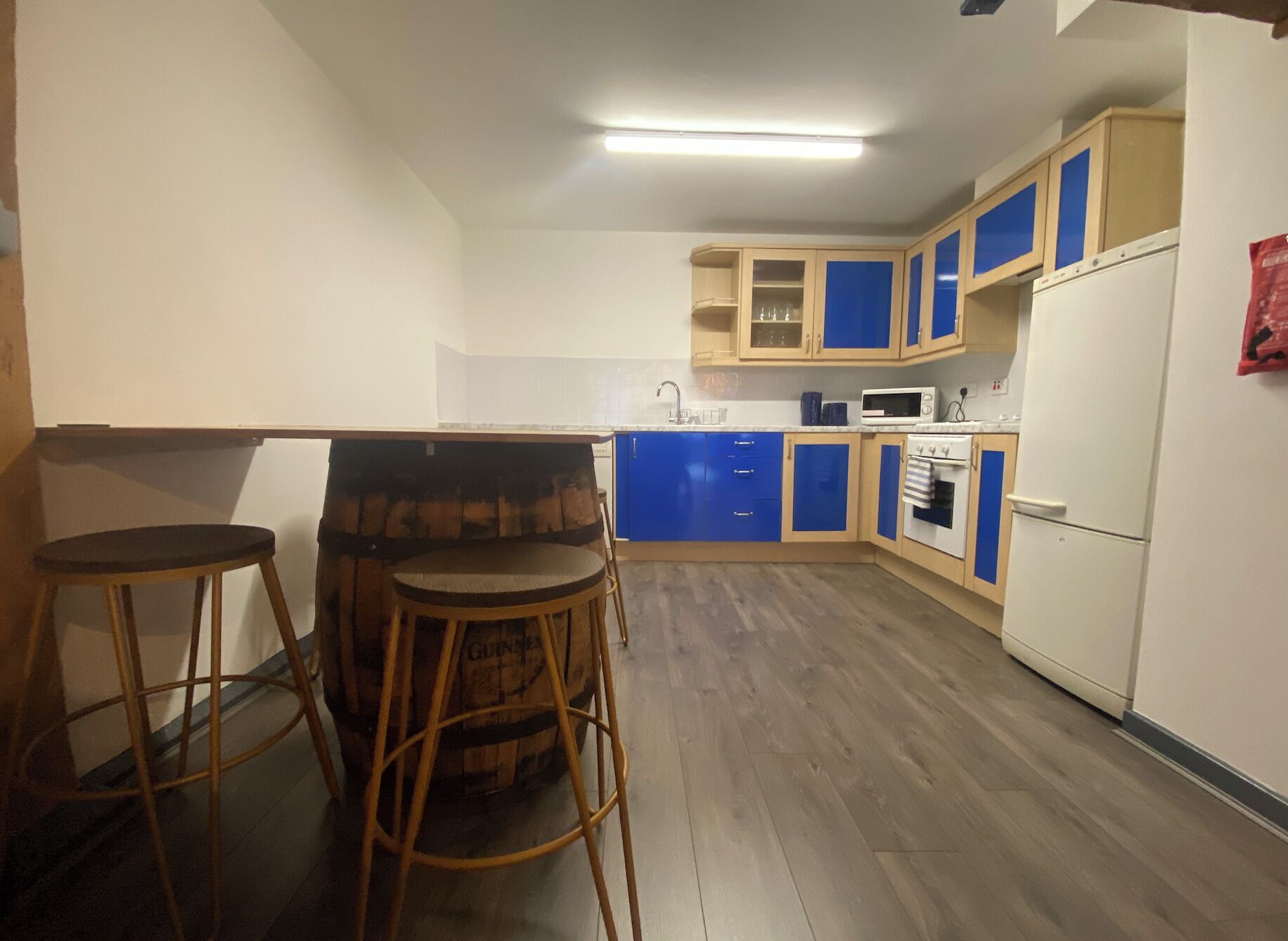 Una cocina compacta con piso de madera, armarios azules y beige, un refrigerador blanco y un pequeño comedor con taburetes.
