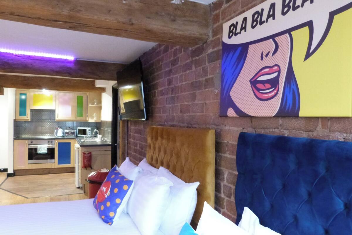 Un dormitorio con un cabecero tapizado en azul, una cama con almohadas blancas y azules, y una cocina visible al fondo.