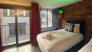 Ein Hotelzimmer mit Bett, einem Fenster mit Blick auf Gebäude, einem roten Vorhang und einer Wandleuchte.