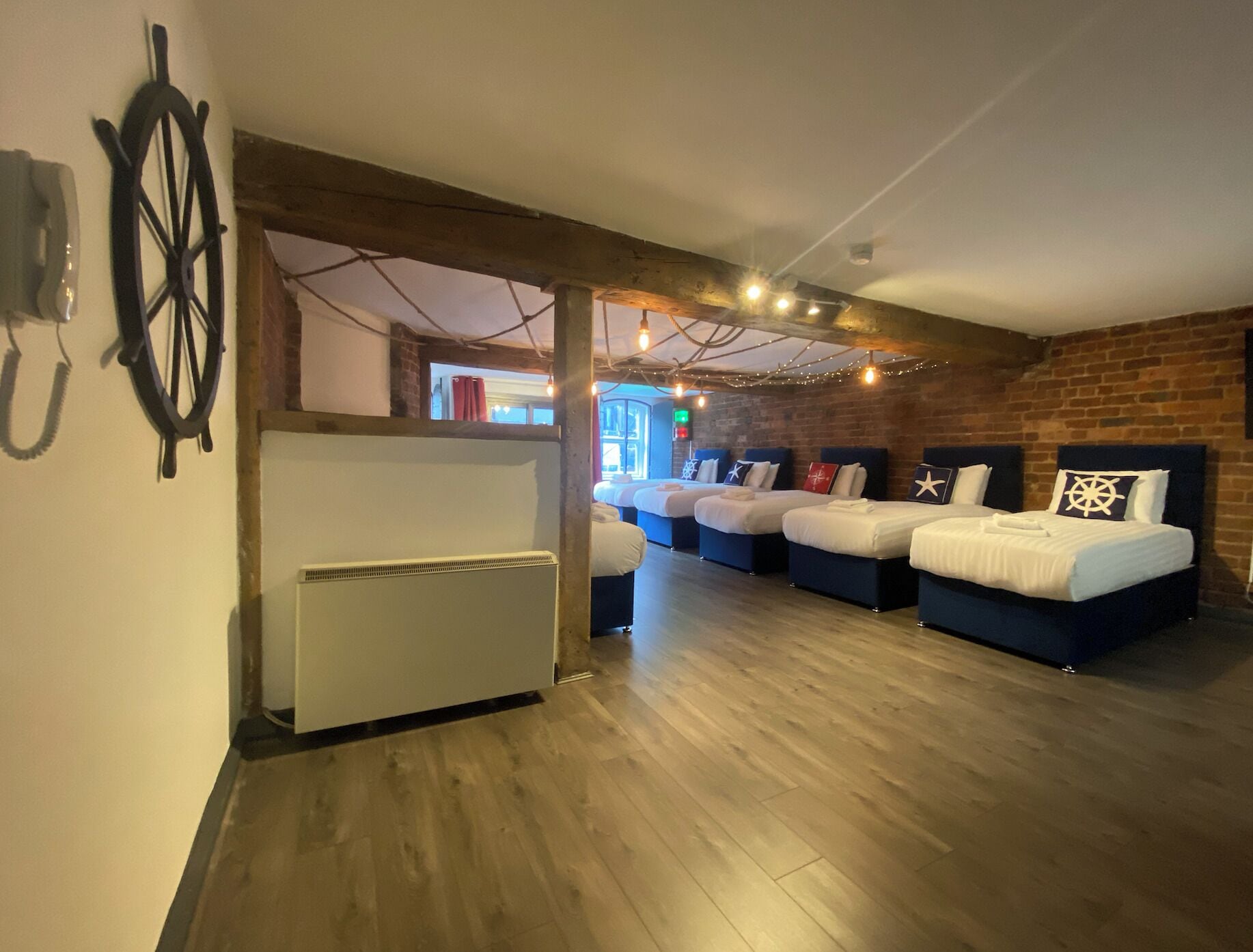 A room with four beds arranged in a row, wooden flooring, a brick wall, and a ceiling with exposed beams and string lights.