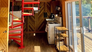A wooden interior with a spiral staircase, a kitchen area, and a large window providing a view of the outdoors. - The Aframe Beach front cottage rental on Manitoulin Island (Sheguiandah)