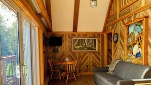 A cozy cabin interior with a wooden floor, a gray sofa, a dining table with chairs, a TV mounted on the wall, and a large window overlooking a deck. - The Aframe Beach front cottage rental on Manitoulin Island (Sheguiandah)
