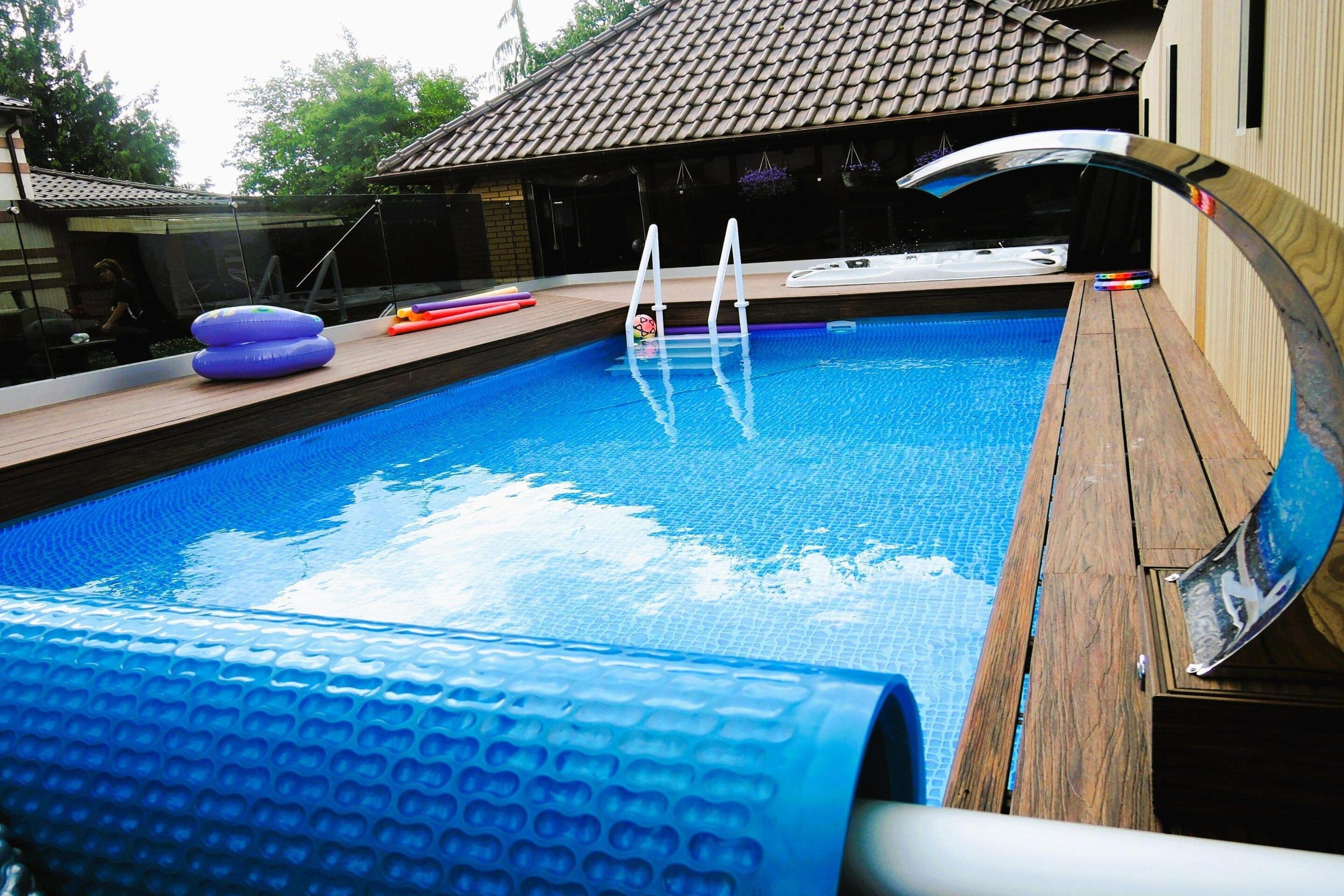 A swimming pool with a wooden deck, a hot tub, and a ladder leading into the pool.