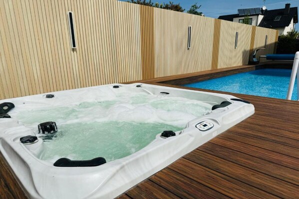 A hot tub with a wooden deck and a pool in the background.