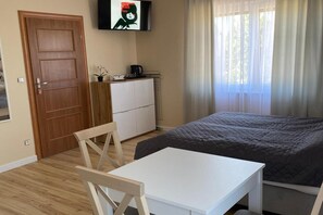 A bedroom with a bed, a white table, a wooden chair, a TV on the wall, and a window with curtains. - Studio in Miedzywodzie Near Baltic Beach (Dziwnów)