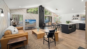 A modern living room with a sofa, coffee table, and TV, a kitchen with dark cabinets and a stove, and large windows with a view of the outdoors.