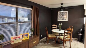 Un coin repas avec une table et des chaises, un jeu de société Monopoly et une fenêtre avec des stores.