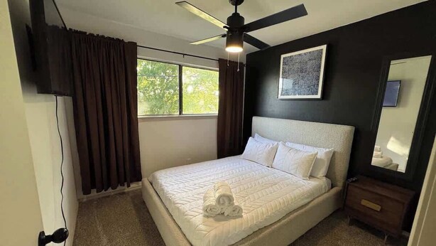 Une chambre à coucher comprenant un lit, un ventilateur de plafond, une fenêtre, une table de chevet et un téléviseur fixé au mur.